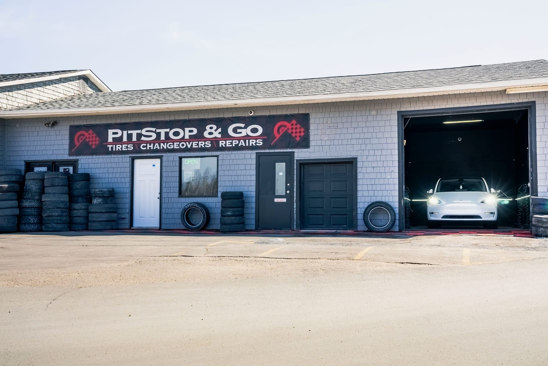 The PitStop & Go storefront on Wilsey Road in daylight, with the shop sign over the doors, stacked tires, and a white Tesla Model Y in the lit service bay